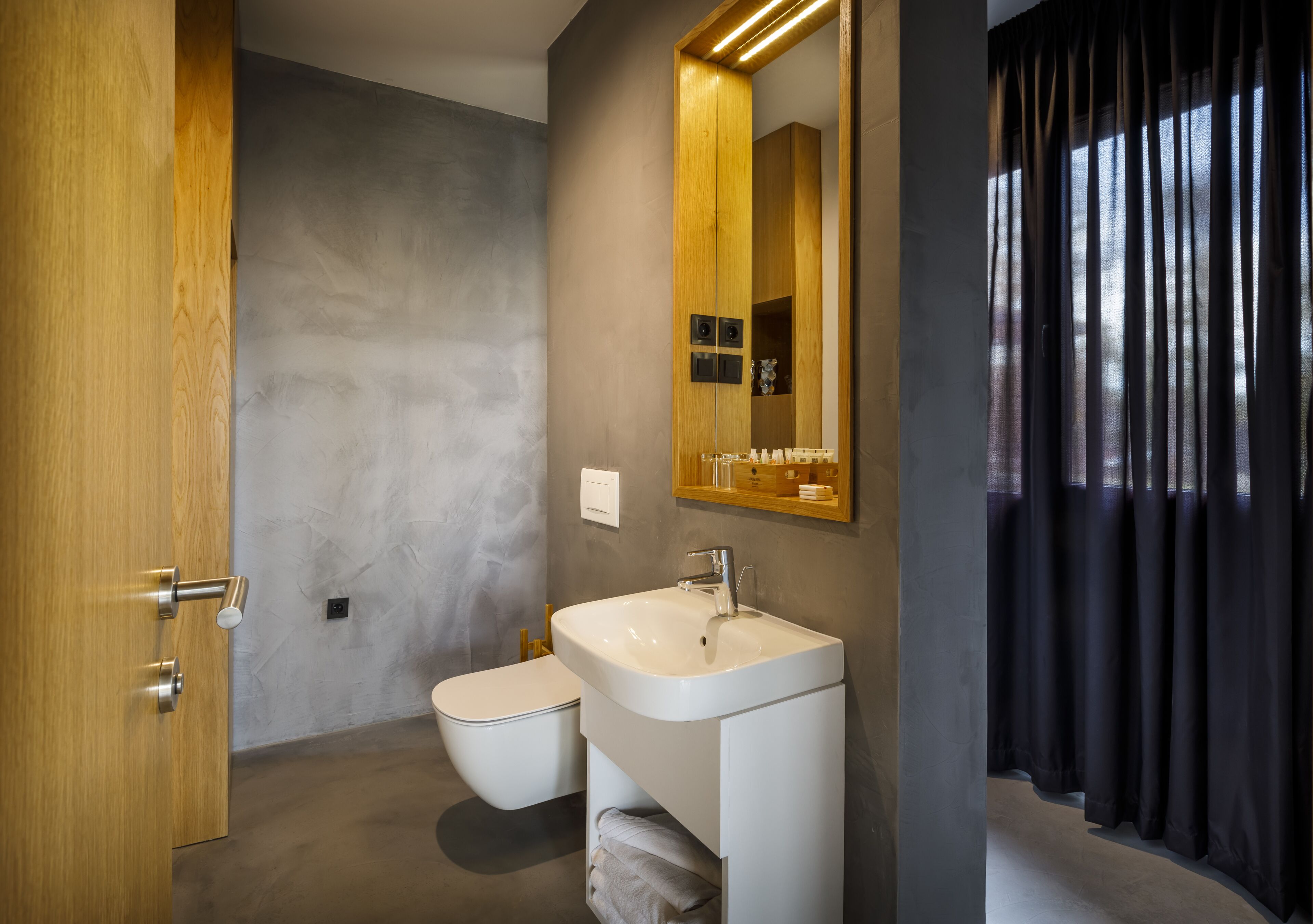 Une salle de bain moderne avec une porte en bois, un lavabo blanc, des toilettes et un miroir surmonté d’une étagère.