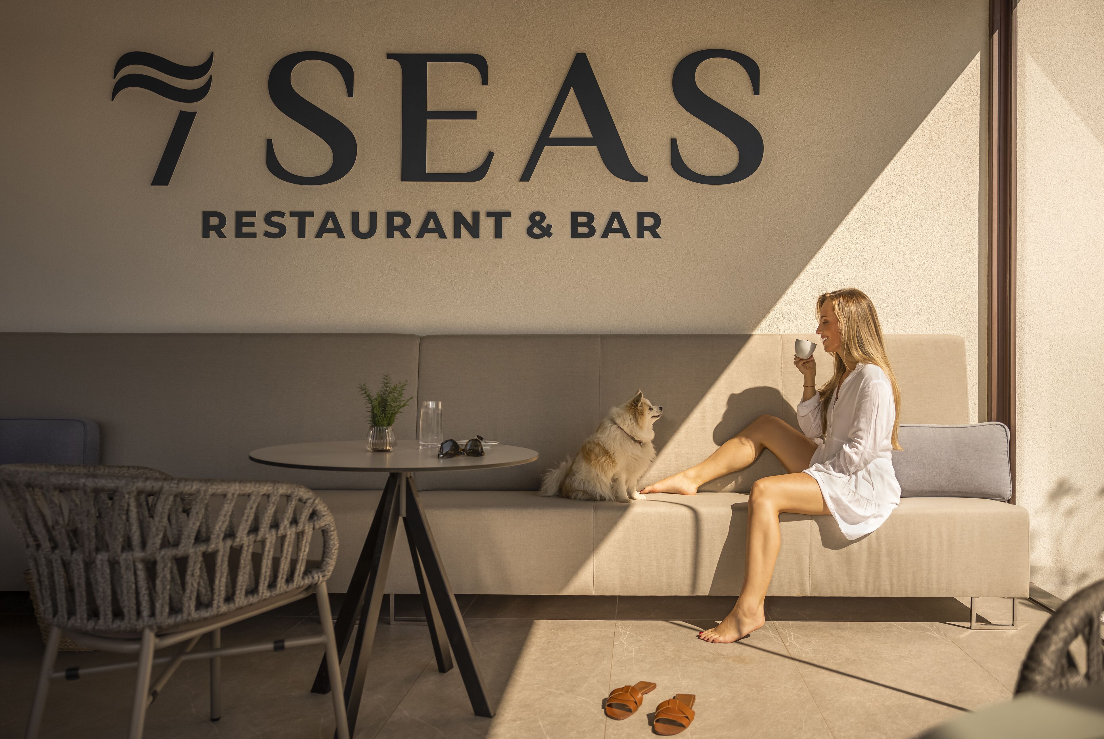 A woman sitting on a sofa in a restaurant with a dog, a table with a plant, and a sign reading "SEAS RESTAURANT & BAR".