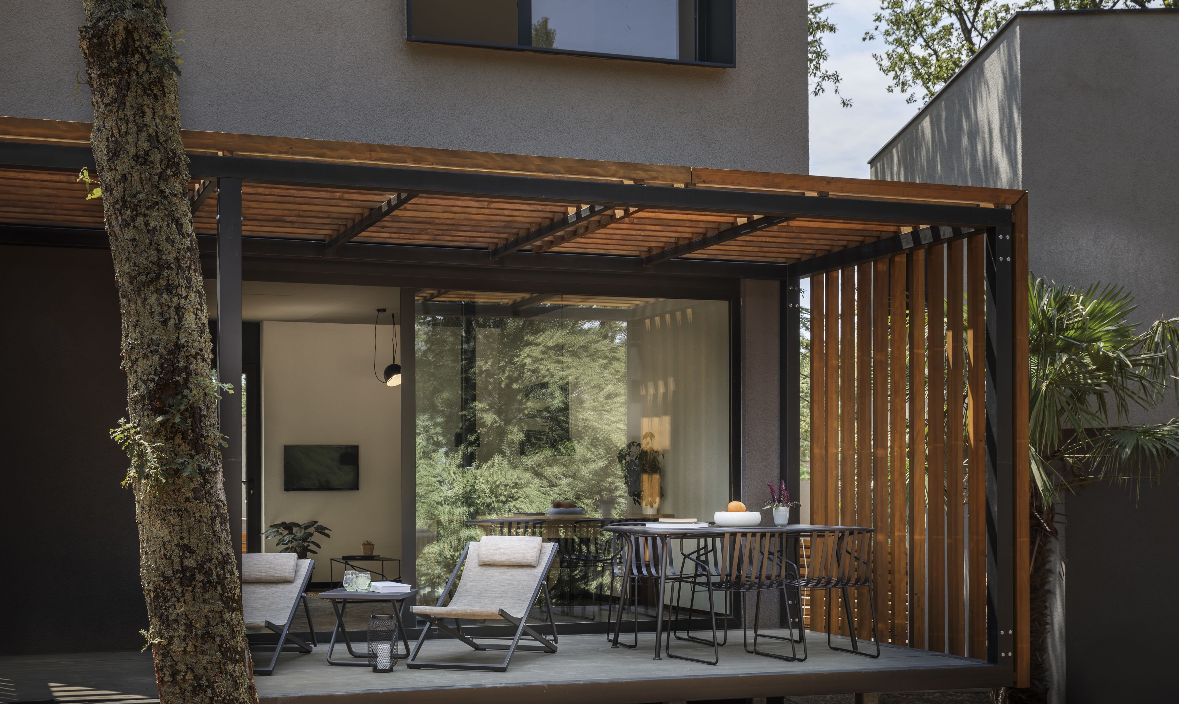 A modern outdoor patio with a glass enclosure, wooden ceiling, and a seating area with chairs and a table.