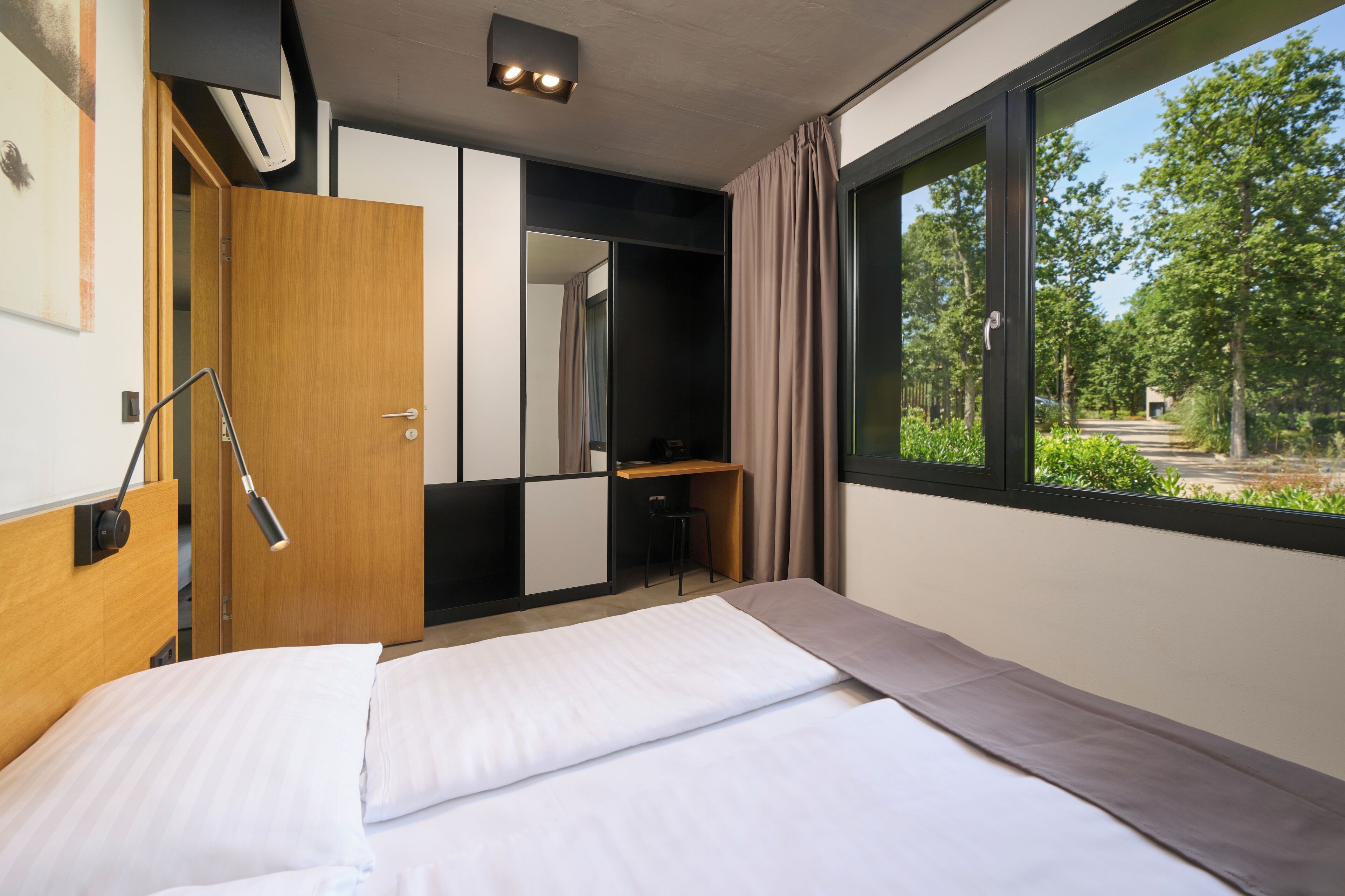 Een moderne hotelkamer met een bed, een bureau, een stoel en een groot raam met uitzicht op groen.
