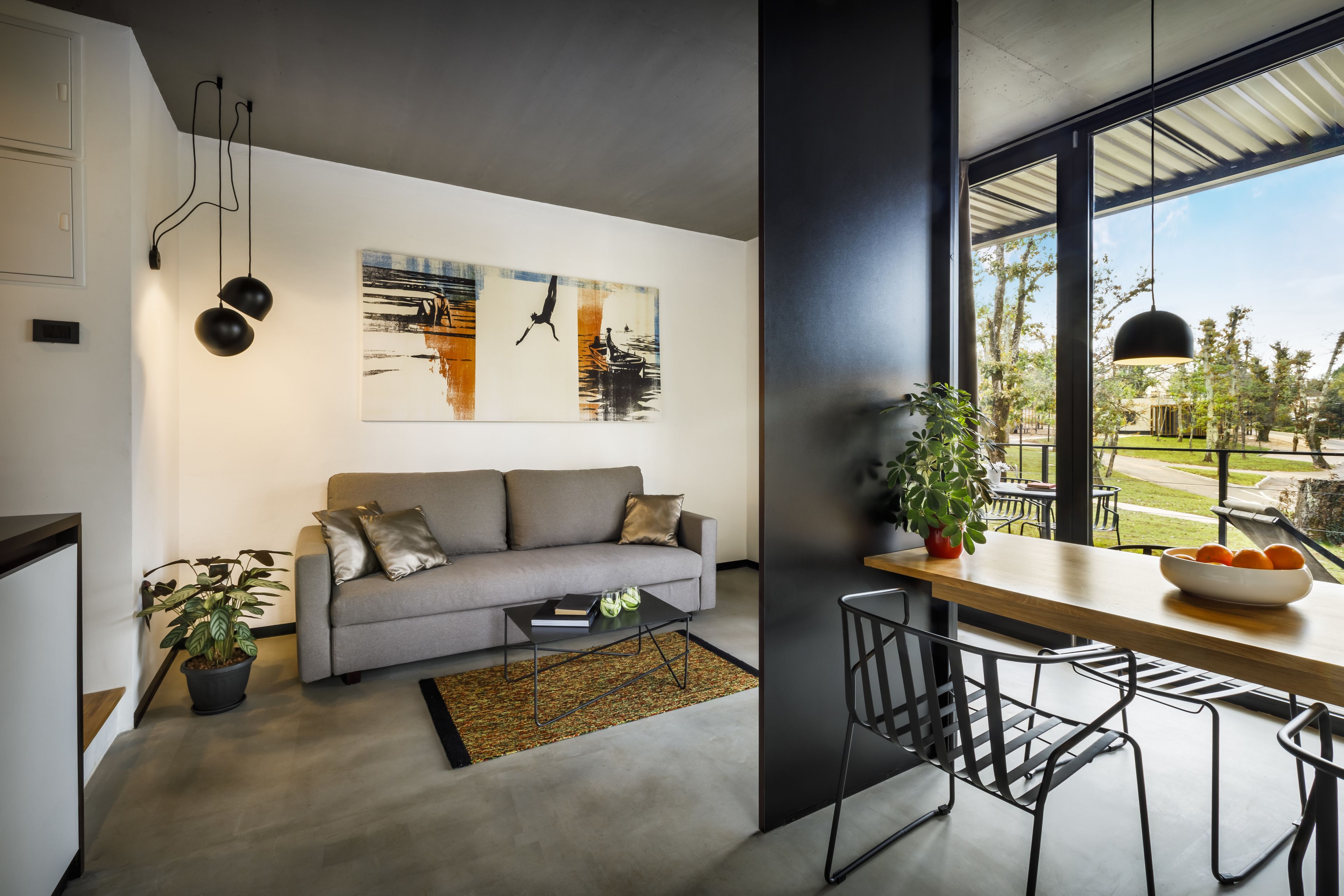 Un salon moderne avec un canapé gris, une table basse et un coin repas comprenant une table et des chaises en bois. Une grande fenêtre offre une vue sur l’extérieur.