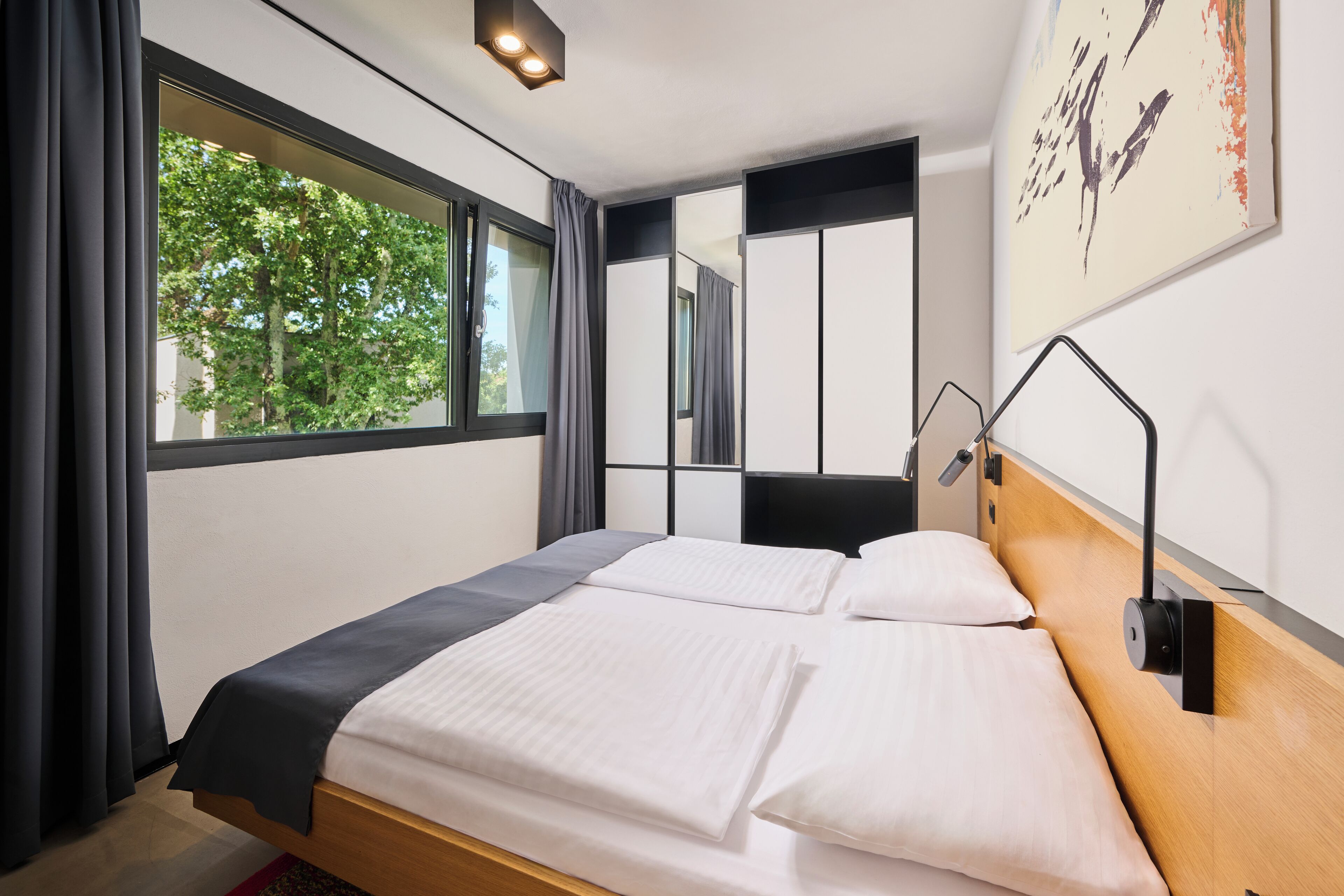 A modern hotel room with a large bed, a bedside table, a mirror, and a window with curtains.