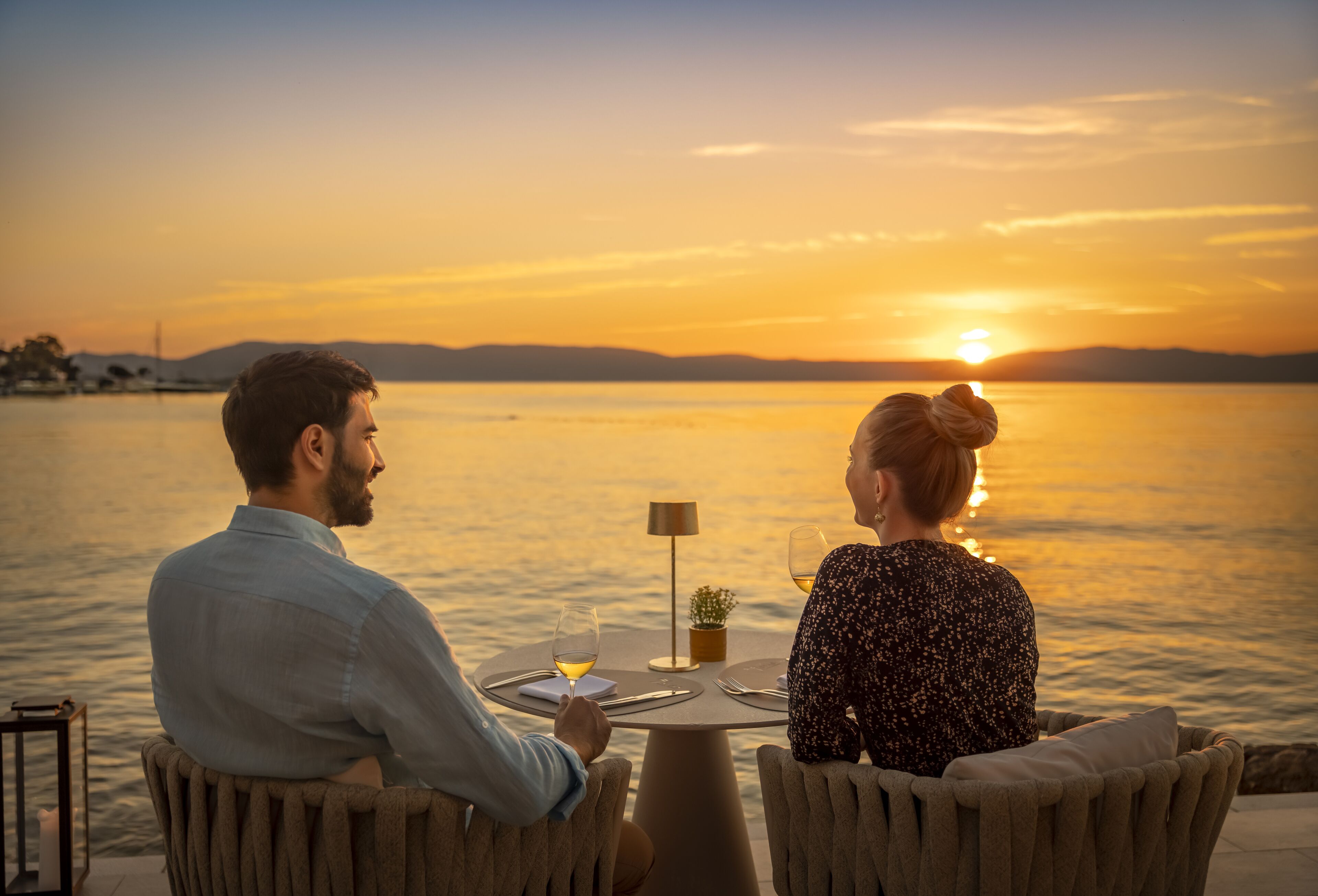 Two people seated at a round table with a view of the sunset over water.