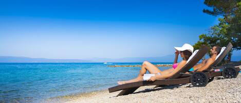 Two people relaxing on wicker lounge chairs by a pebble beach with a clear view of the sea and distant mountains.