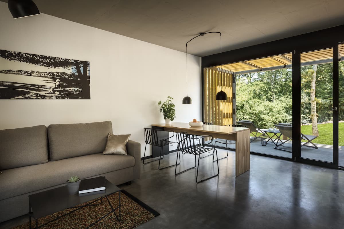 Una sala de estar moderna con sofá, una mesa de centro y un comedor. Se ve un ventanal con vistas a la vegetación.