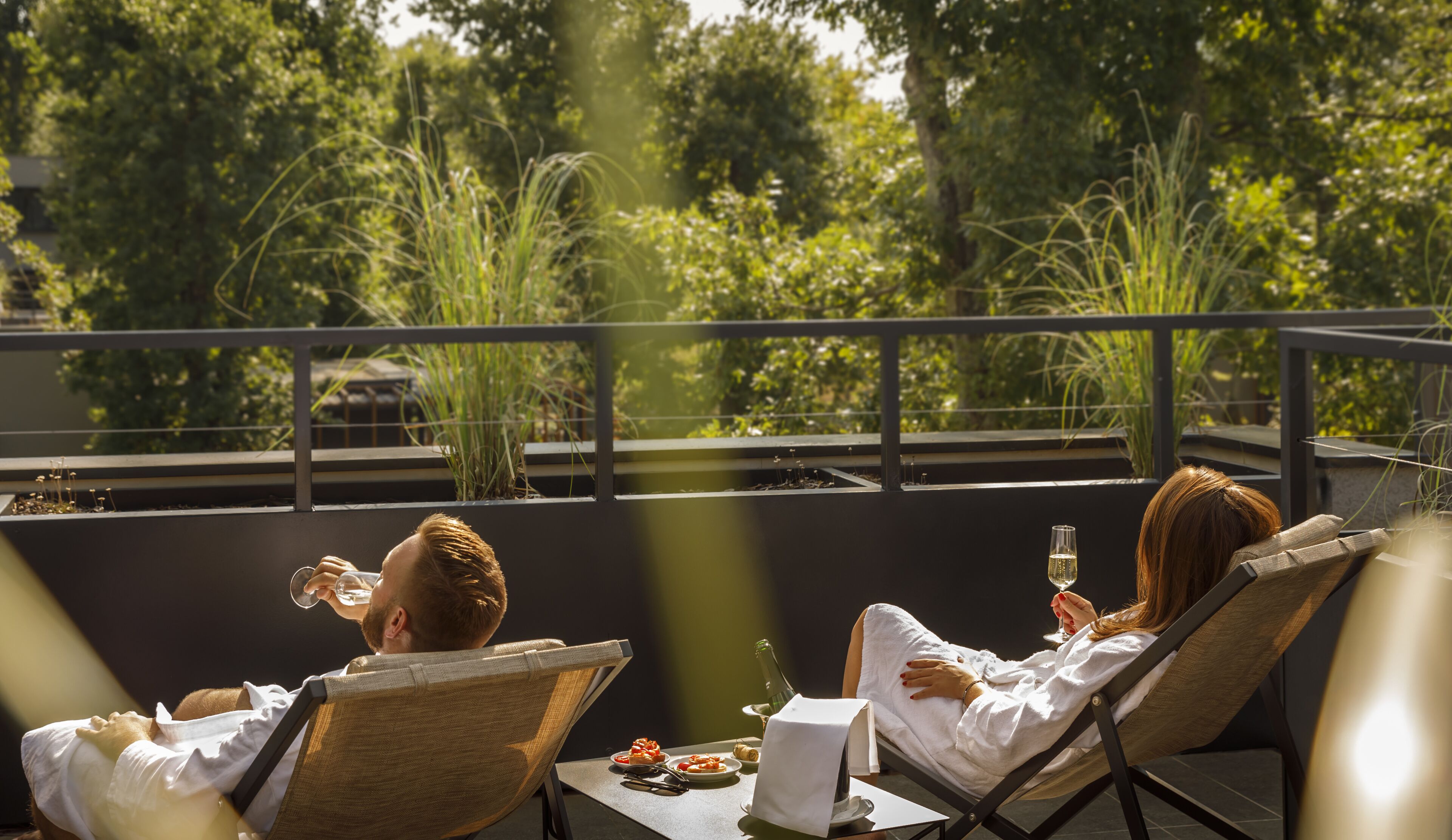 Two people relaxing on a balcony with a table set for a meal, overlooking a garden.