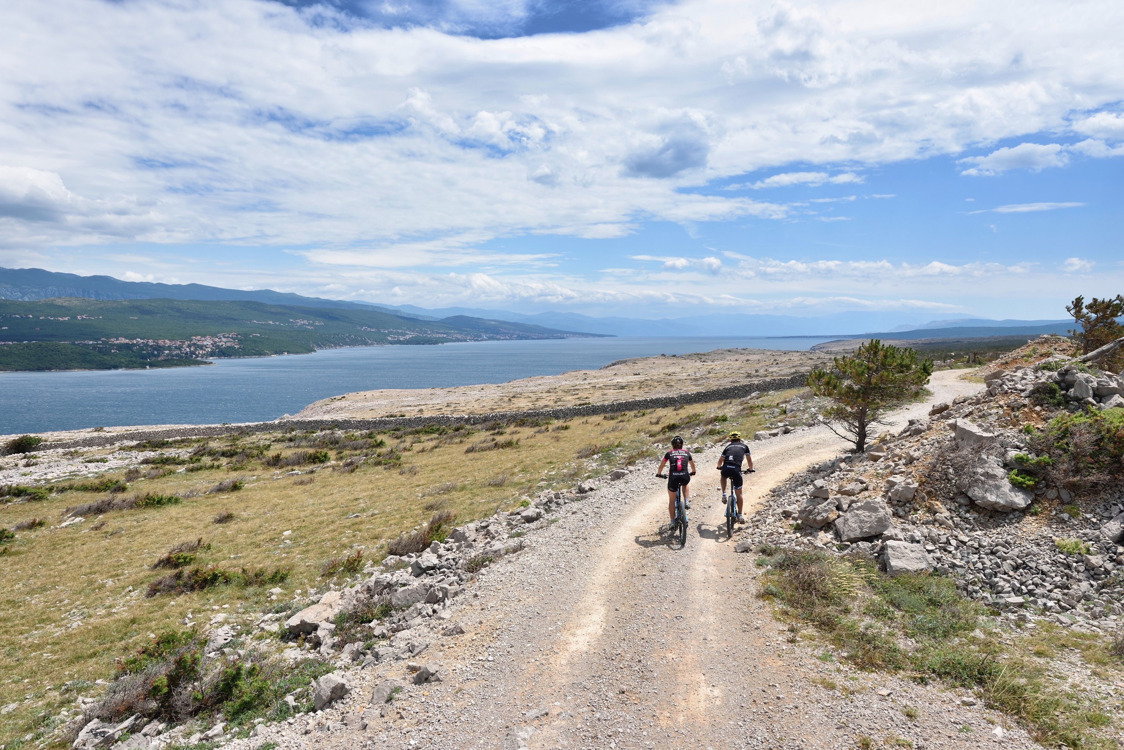 Un chemin de terre avec deux cyclistes, un terrain rocailleux et une étendue d’eau au loin, sous un ciel partiellement nuageux.