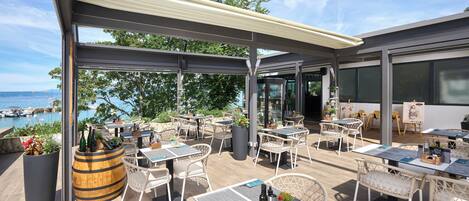 An outdoor patio with tables and chairs, a wooden barrel, and a view of the sea and boats.