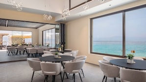Een moderne eetruimte met ronde tafels en stoelen, grote ramen met uitzicht op zee, en decoratieve hanglampen.