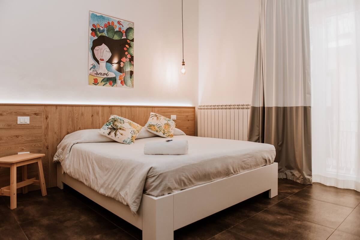 Un dormitorio con cabecera de madera, una cama con almohadas estampadas, una mesita de noche y una lámpara de pared.