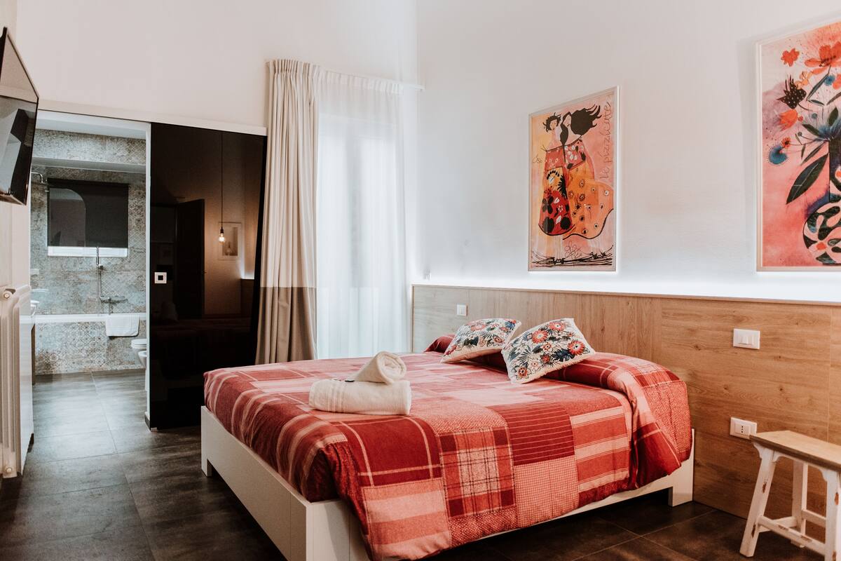 Un dormitorio con cama, cabecero de madera, un taburete pequeño de madera y un baño visible a través de una puerta abierta.