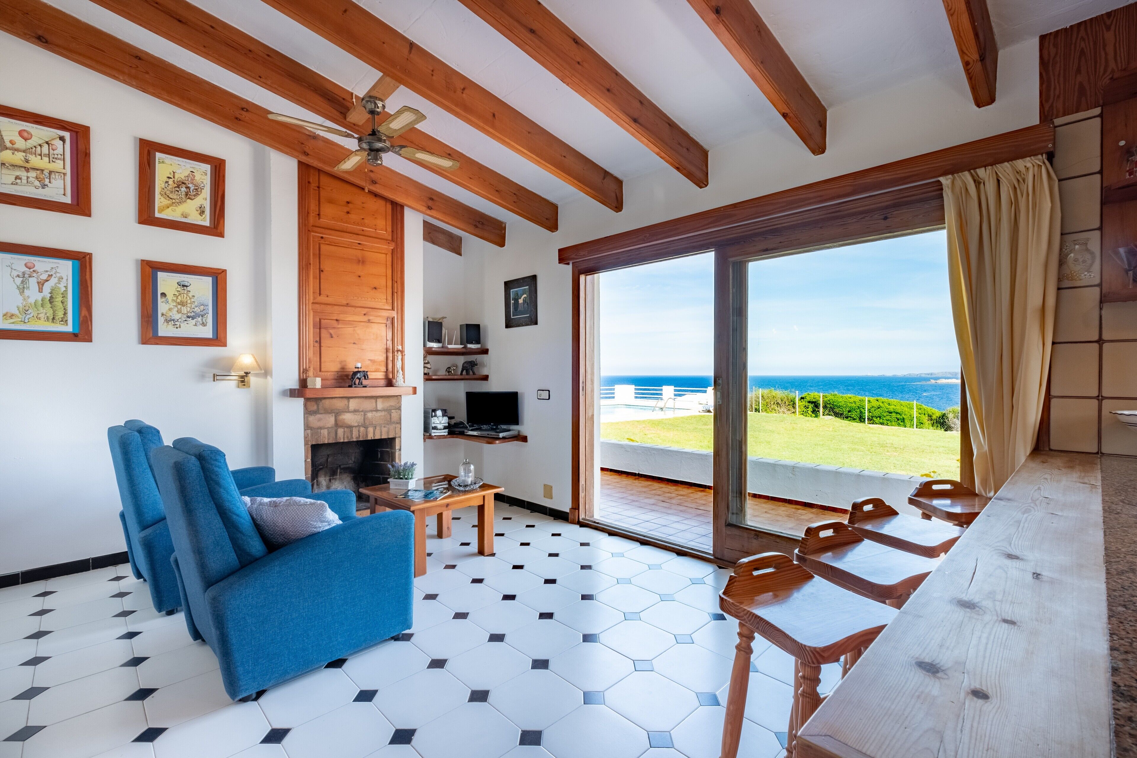 A living room with a blue sofa, a fireplace, a TV, and a view of the sea through a glass door.