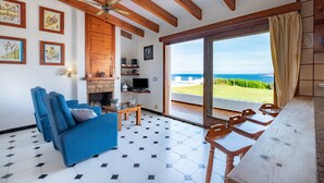 A living room with a blue sofa, a fireplace, a TV, and a view of the sea through a glass door. - Directly by the sea with greenhouse - Villa Solivent (Mercadal)
