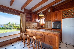 Une cuisine avec des meubles de rangement en bois, un coin bar avec des tabourets, un réfrigérateur et une vue sur l’extérieur.
