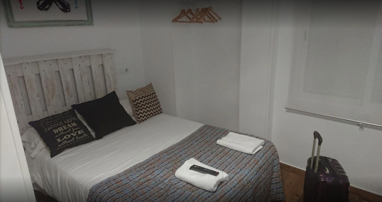 A neatly made bed with a patterned bedspread, a pillow with a motivational quote, and a remote control on the bed. There is a suitcase on the floor to the right.