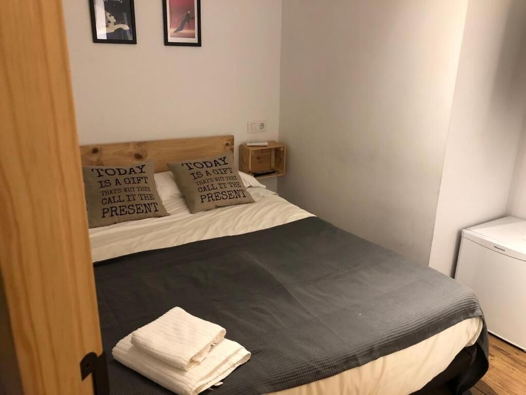 Ein ordentlich bezogenes Bett mit einer schwarz-weißen Tagesdecke, zwei Kissen mit motivierenden Sprüchen, ein kleiner Nachttisch mit Lampe und eine weiße Wand mit zwei gerahmten Bildern.