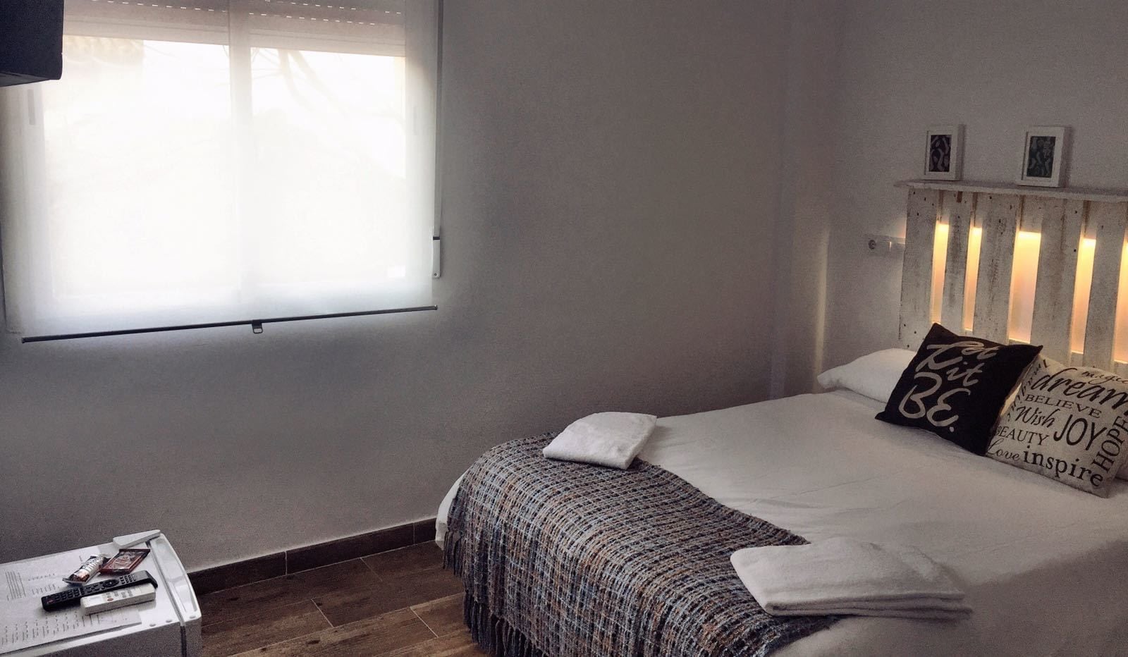 A bedroom with a bed, a bedside table with a remote control, and a window with blinds.