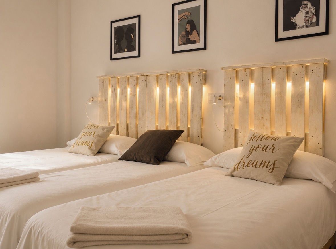 Une chambre d’hôtel avec deux lits, chacun doté d’une tête de lit ornée de lattes verticales et d’un éclairage intégré. On y trouve des coussins décoratifs avec des motifs textuels, et la chambre présente un design minimaliste.
