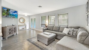 A spacious living room with a grey sectional sofa, a coffee table, and a TV mounted on the wall. There are two windows with blinds, a door with glass panels, and a small round mirror on the wall. - Canal fun! Boat, Pool, Hot tub, Bikes & Kayaks (Cape Coral)