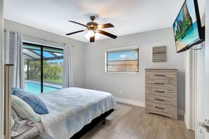 Une chambre à coucher avec un lit, un ventilateur de plafond, une fenêtre avec des stores, une commode en bois et un téléviseur fixé au mur.