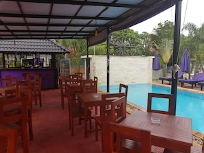 A covered outdoor area with wooden tables and chairs, a pool, and a bar with a drink dispenser. - White Lotus Resort (Siem Reap)
