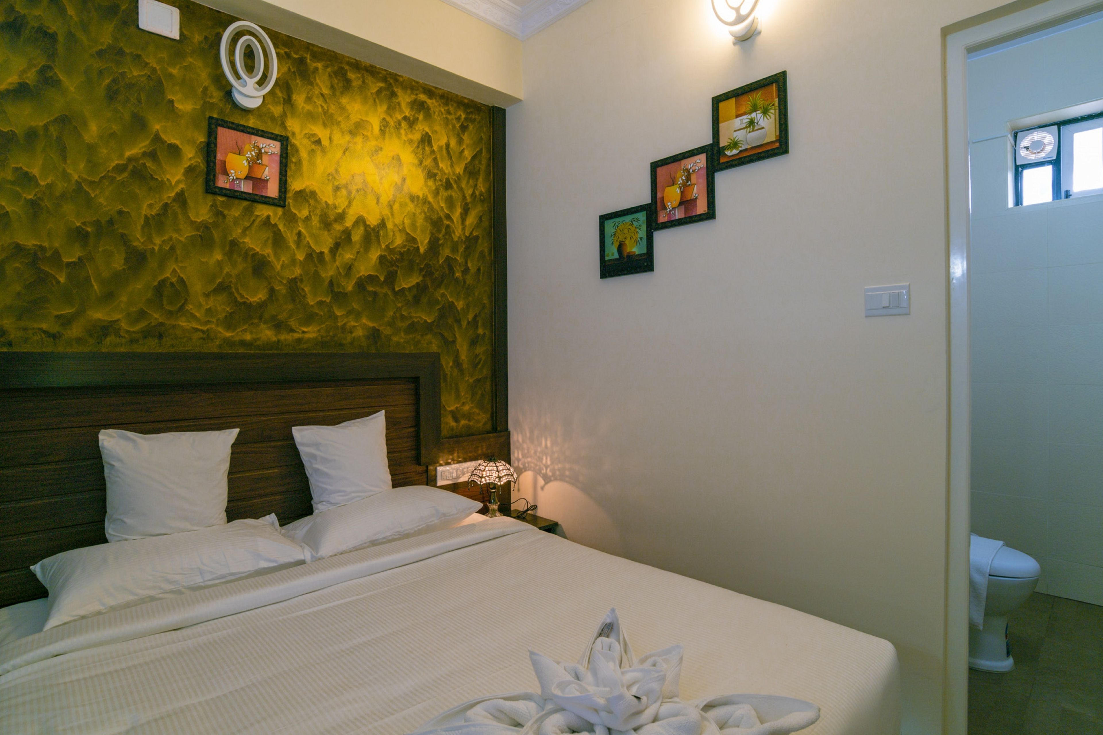 Una habitación de hotel con cama, mesita de noche, lámpara y pared decorada con papel tapiz texturizado y cuadros.