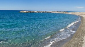 Een strand met helderblauw water en een rotsachtige landtong in de verte.