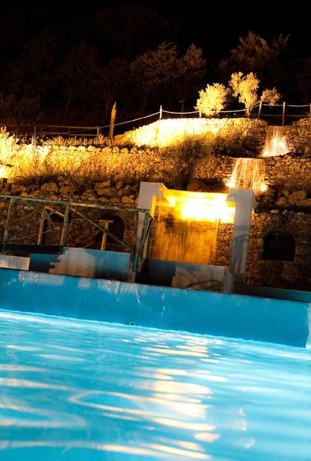 Une piscine agrémentée d’une cascade, entourée d’un mur de pierre et d’une clôture en bois.