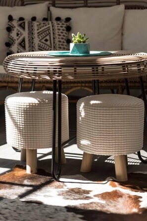 A round wicker table with a teal plant, two white poufs, and a cowhide rug. - Kumba Hostel (Chania)