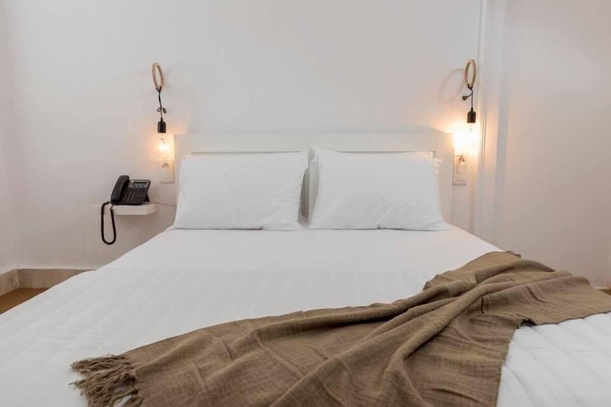 Cama com lençóis brancos, manta marrom e duas luminárias laterais.