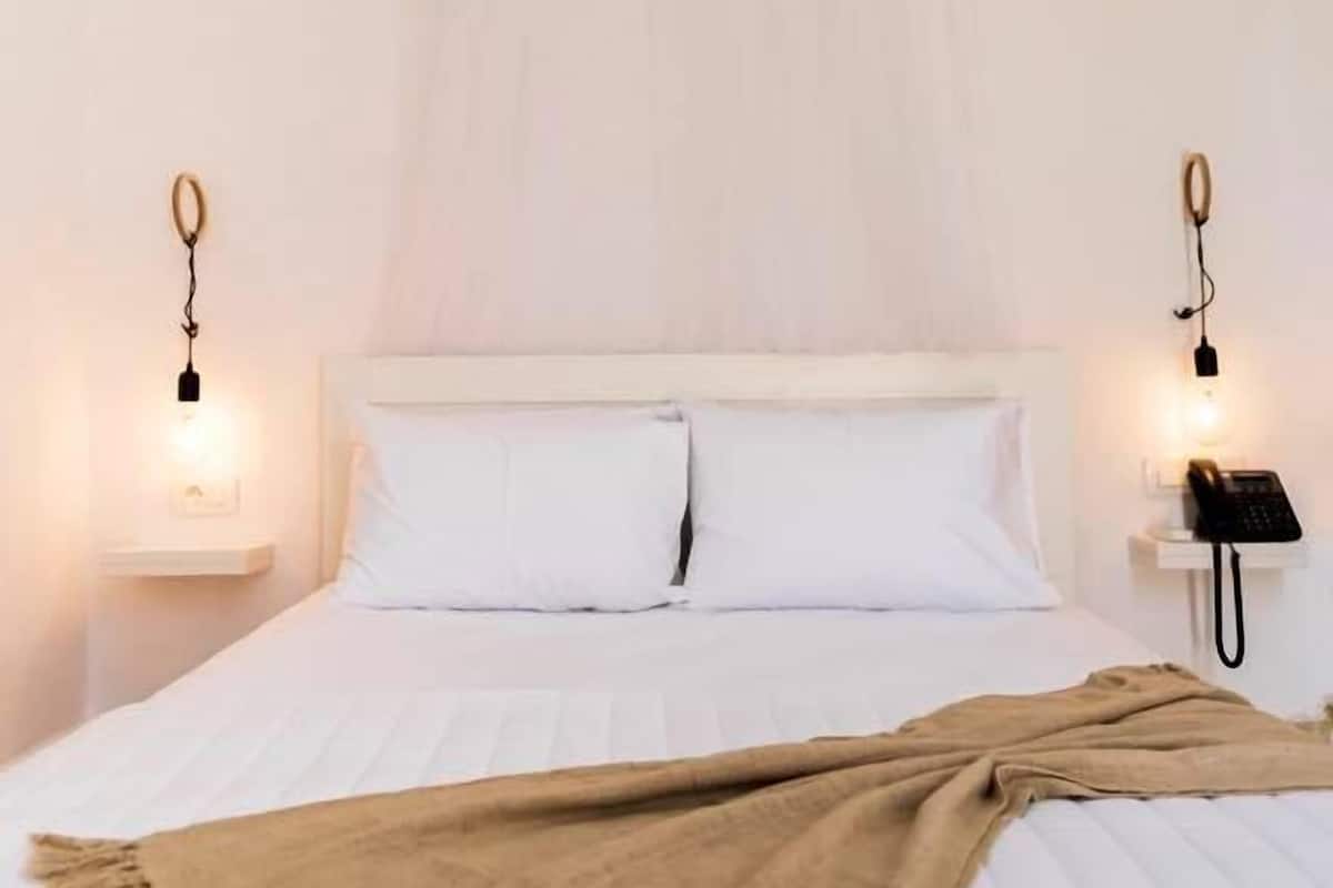 Cama com roupa de cama branca e manta bege, duas mesas de cabeceira com luminárias e um telefone, e uma parede branca.