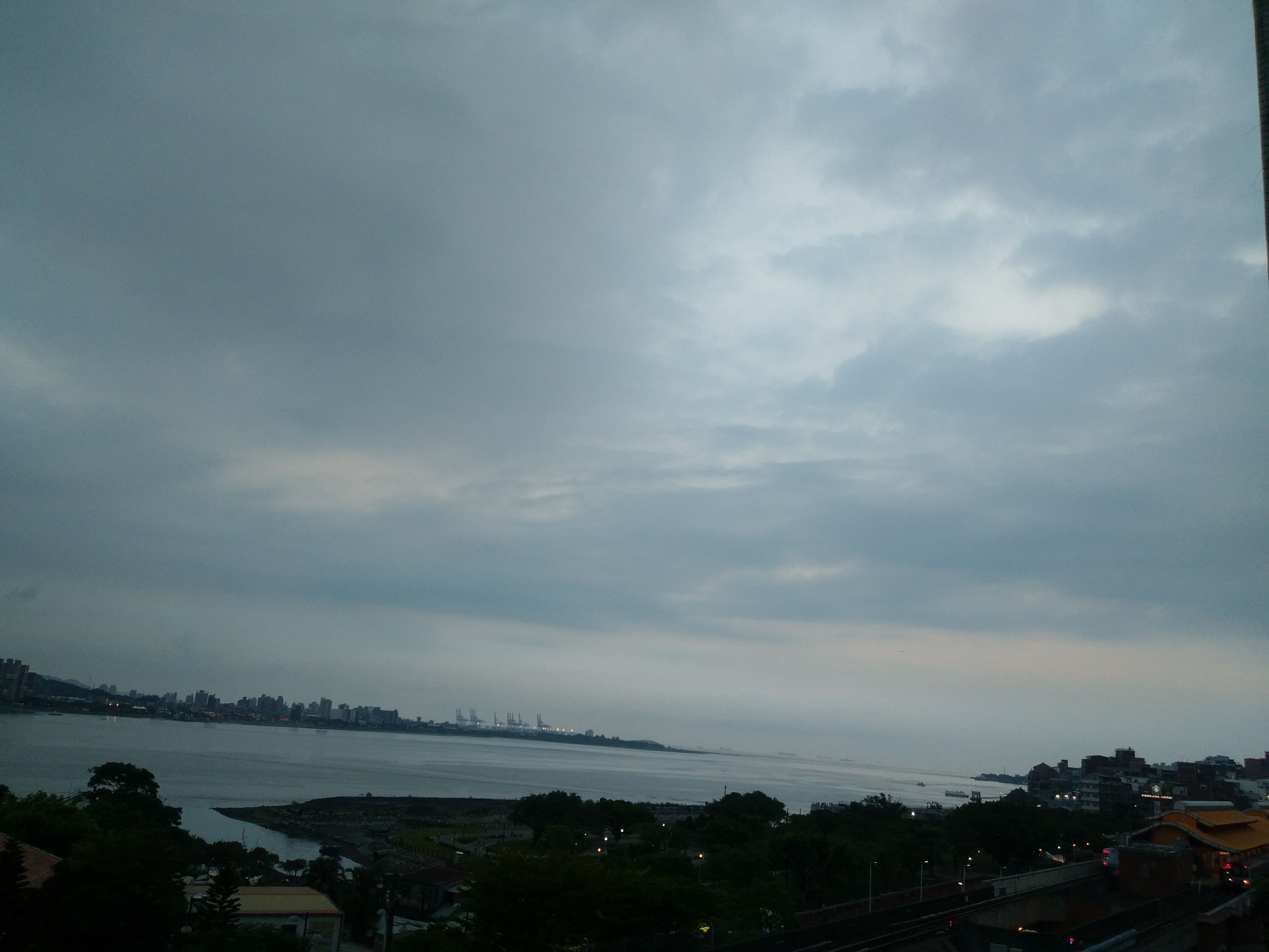 A coastal cityscape with a view of the sea, buildings, and a bridge in the distance.