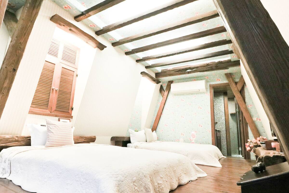 A bedroom with two beds, wooden beams, a window with shutters, and a floral-patterned wall.