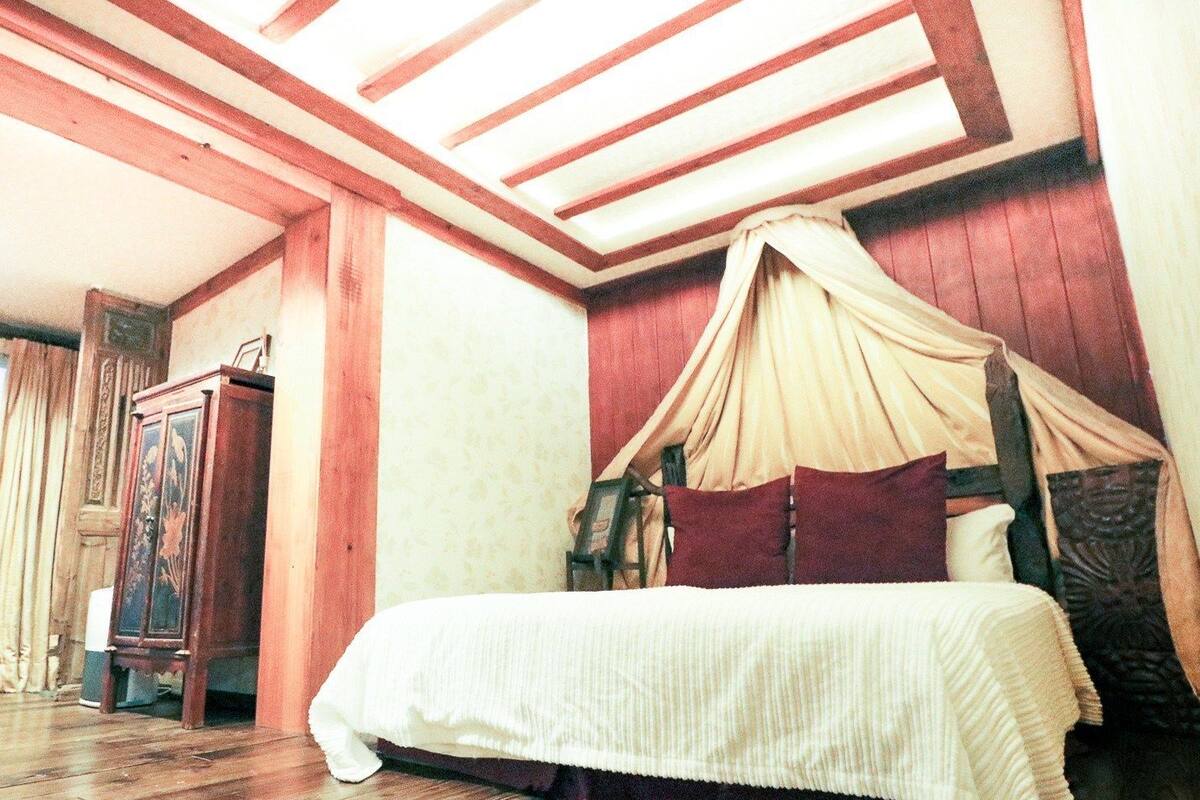 A bedroom with a canopy bed, wooden flooring, a wardrobe, and a large window.