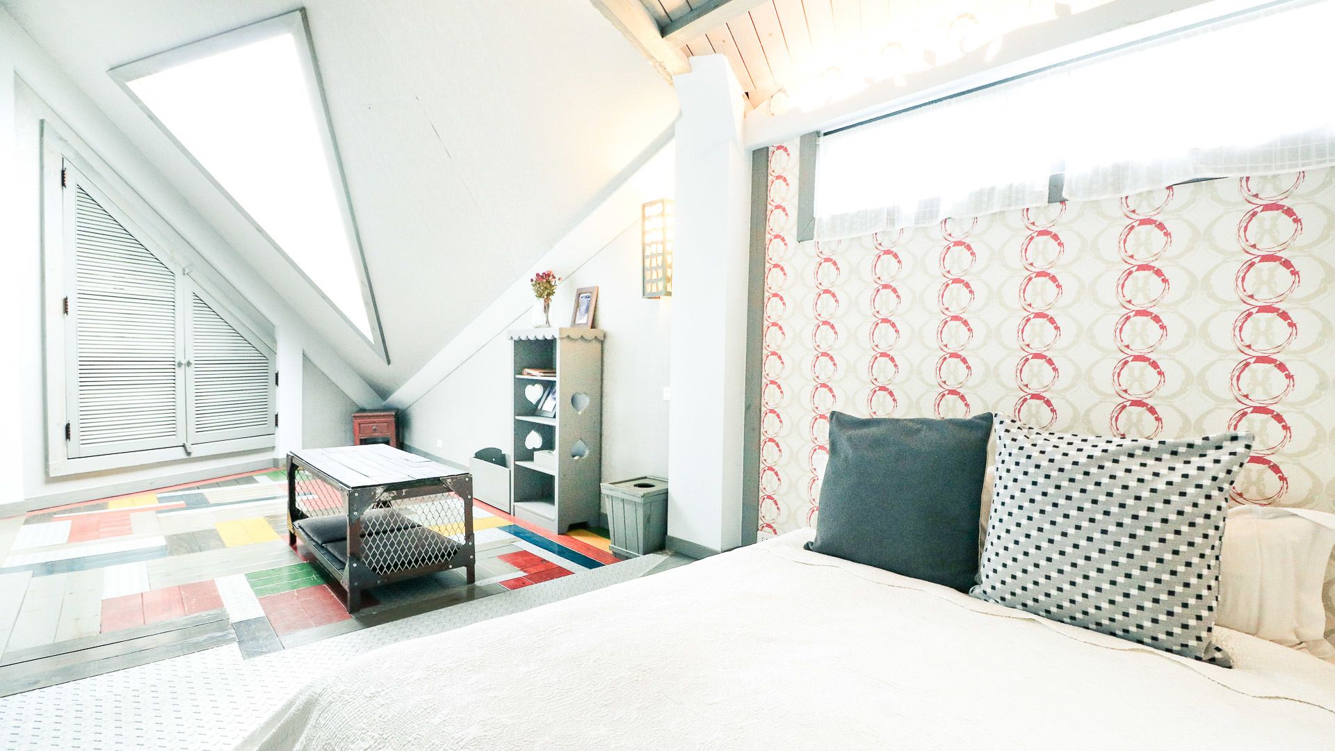 A bedroom with a bed, a small table, a bookshelf, a window, and a patterned curtain.