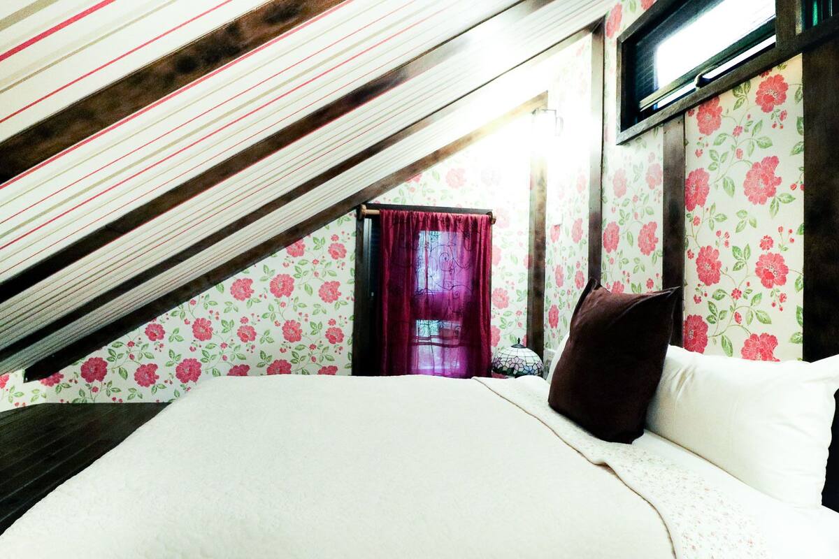 A bedroom with a sloped ceiling, floral wallpaper, a bed with white bedding, a dark brown pillow, and a window with a curtain.