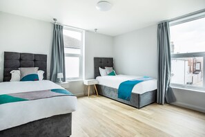 A modern hotel room with two beds, a window with curtains, a bedside table with a lamp, and a view of the city.