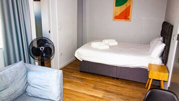 Een hotelkamer met een bed, een stoel, een klein houten tafeltje en een ventilator.