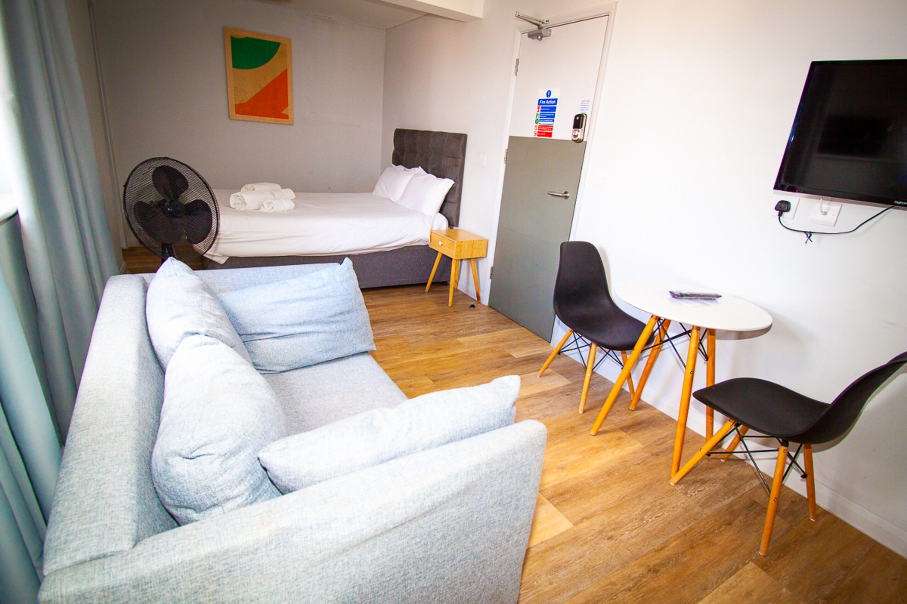 A hotel room with a bed, a sofa, a small table with chairs, a fan, and a TV mounted on the wall.