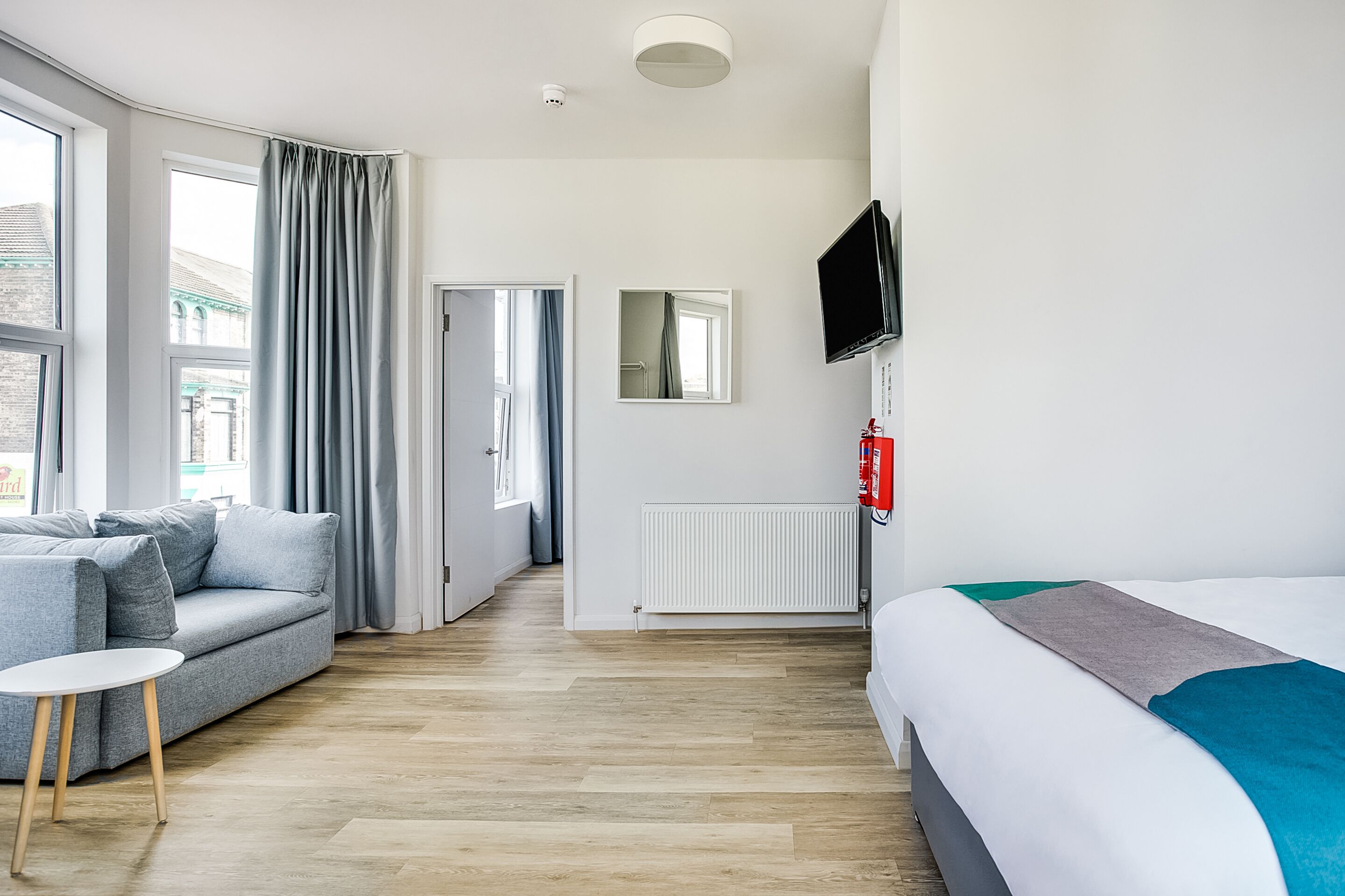 Et moderne hotelværelse med en grå sofa, et lille rundt bord, en seng med et blågråt og gråt tæppe, et vægmonteret tv og et vindue med grå gardiner.