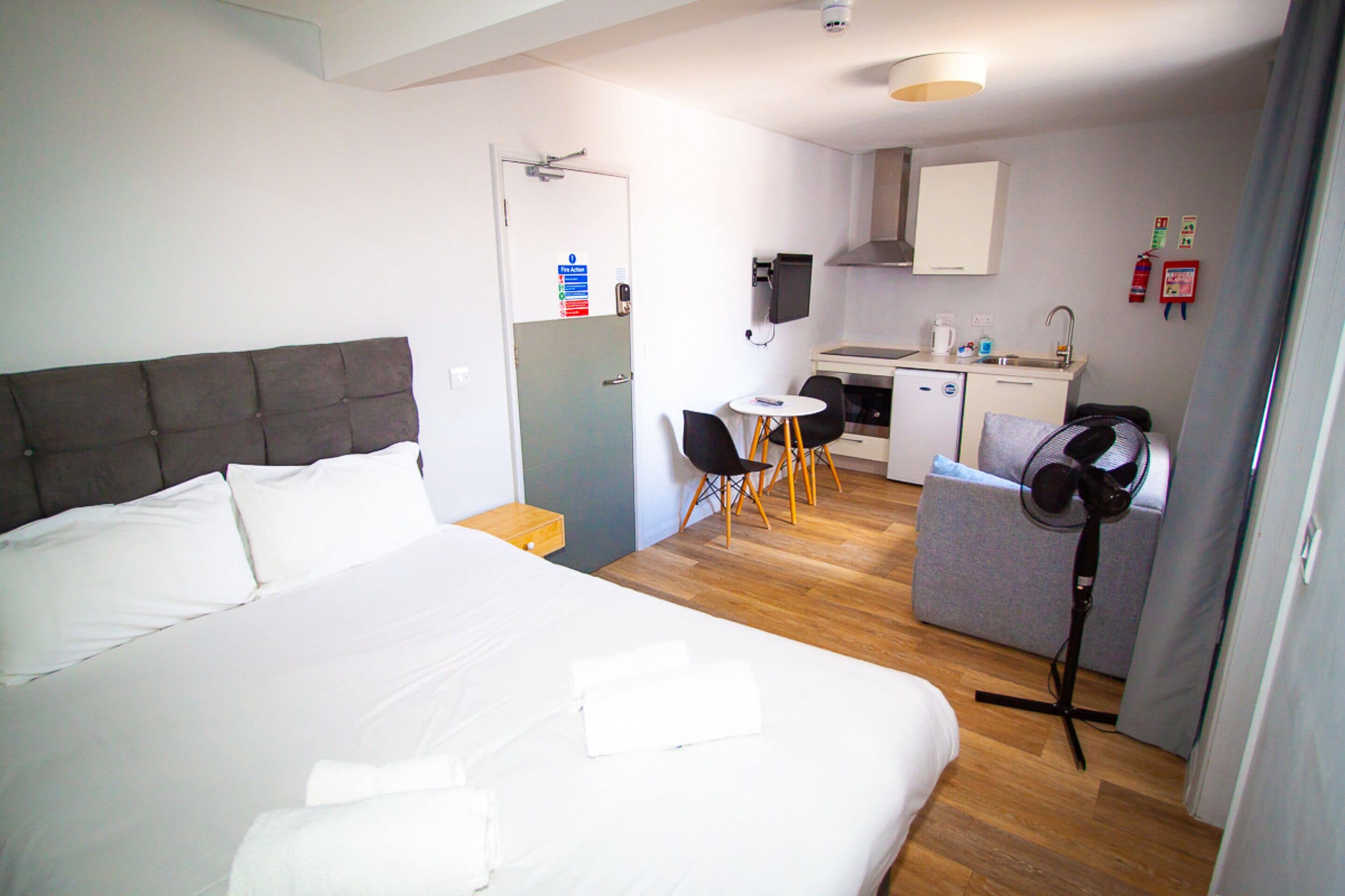 A hotel room with a large bed, a small kitchenette, a dining area, and a fan.