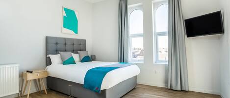 A modern bedroom with a grey upholstered bed, a wooden bedside table, a mounted TV, and a large window with curtains.