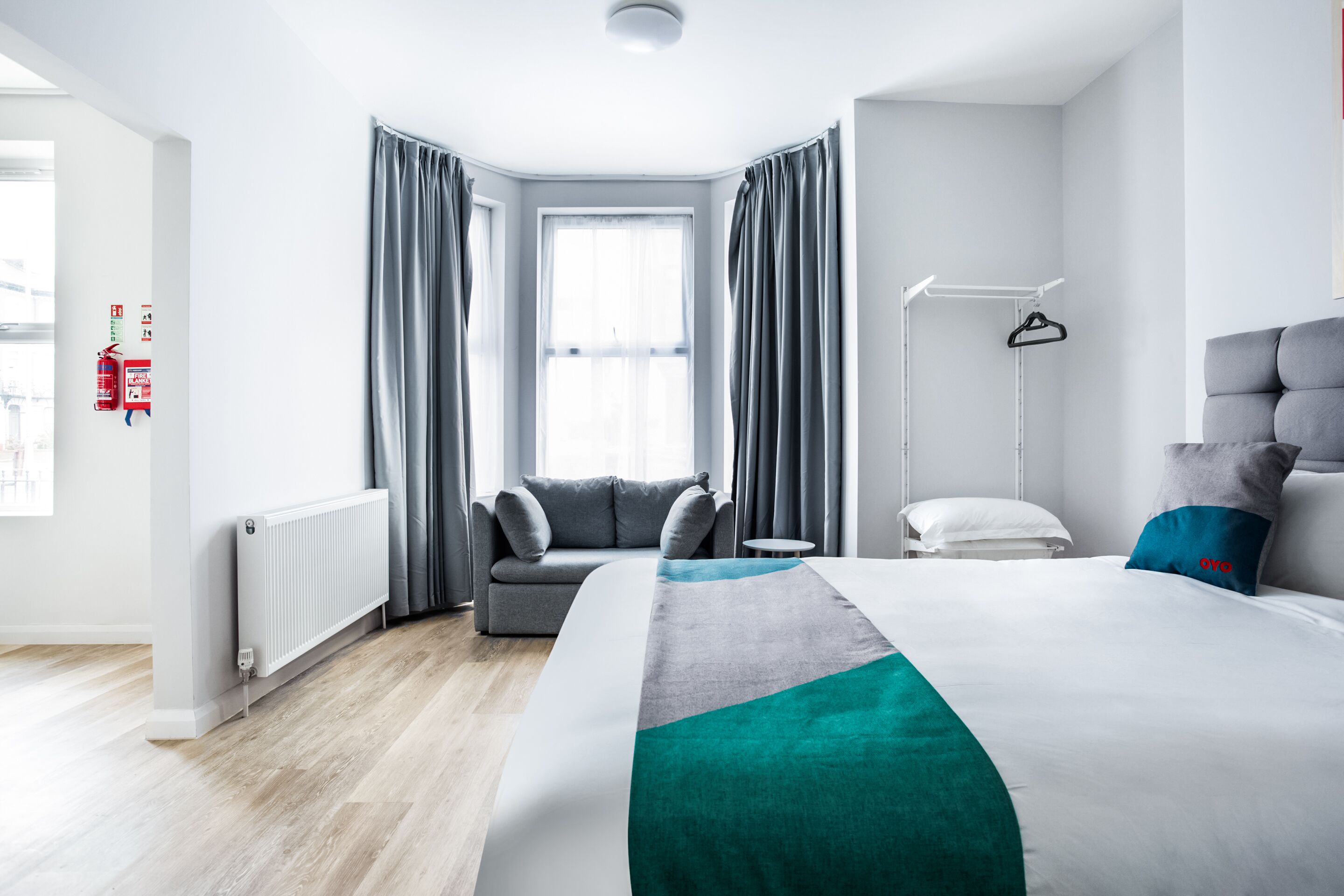 Een moderne hotelkamer met een groot bed, grijze gordijnen, een grijze bank, een kleine tafel, een witte stoel en een radiator.