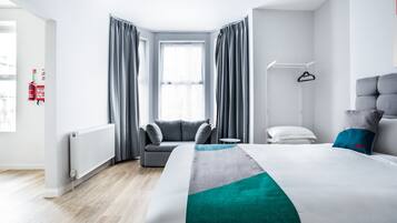 A modern hotel room with a large bed, grey curtains, a grey sofa, a small table, a white chair, and a radiator.