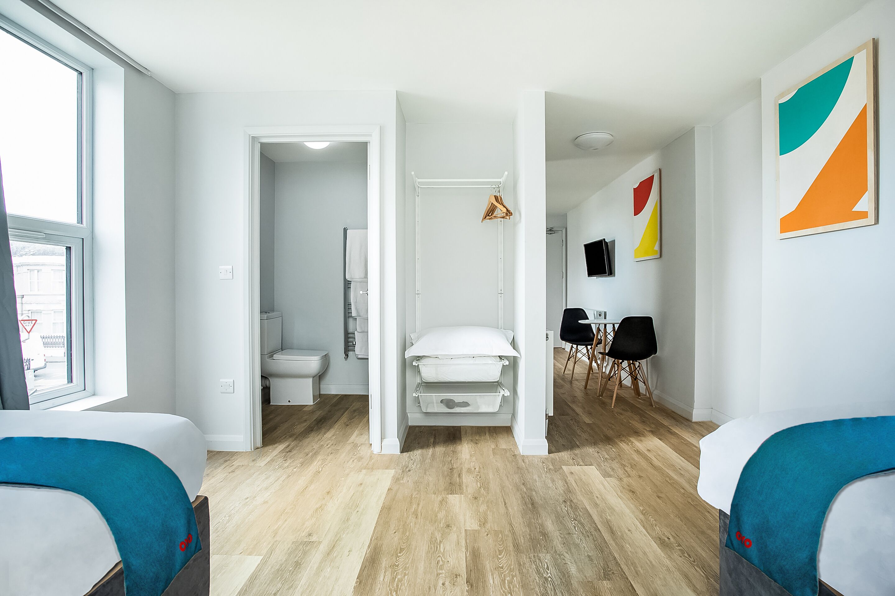 A modern hotel room with a wooden floor, a bed with a blue headboard, a white wardrobe, and a bathroom visible through an open door.