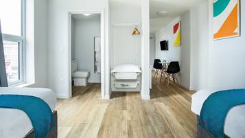 A modern hotel room with a wooden floor, a bed with a blue headboard, a white wardrobe, and a bathroom visible through an open door.