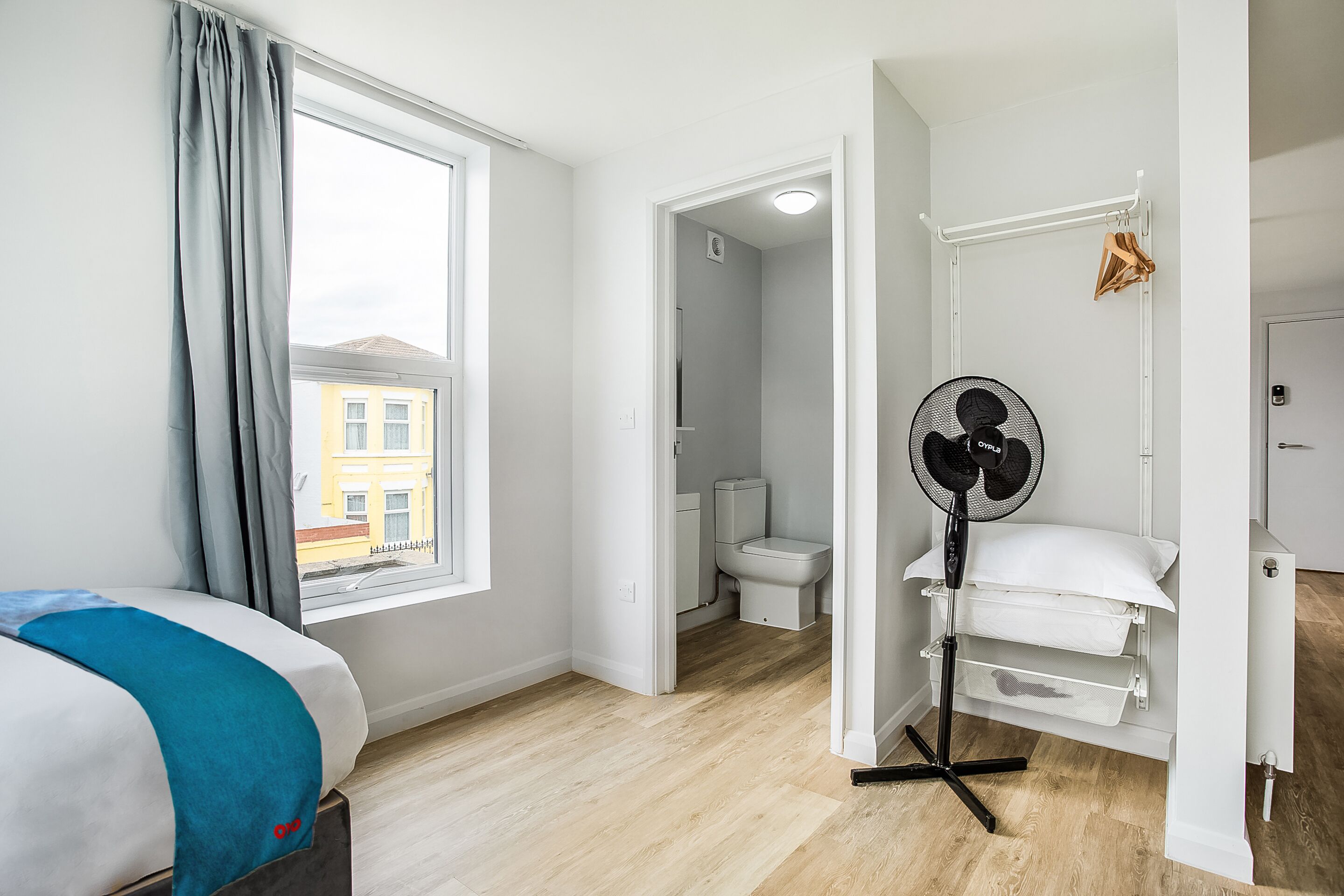 Un dormitorio con cama, ventana, ventilador y vista a un edificio.