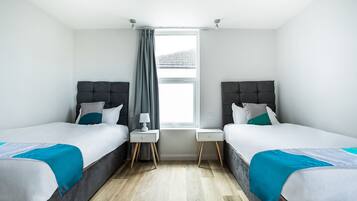A modern hotel room with two beds, a window with curtains, a bedside table with a lamp, and a wooden floor.