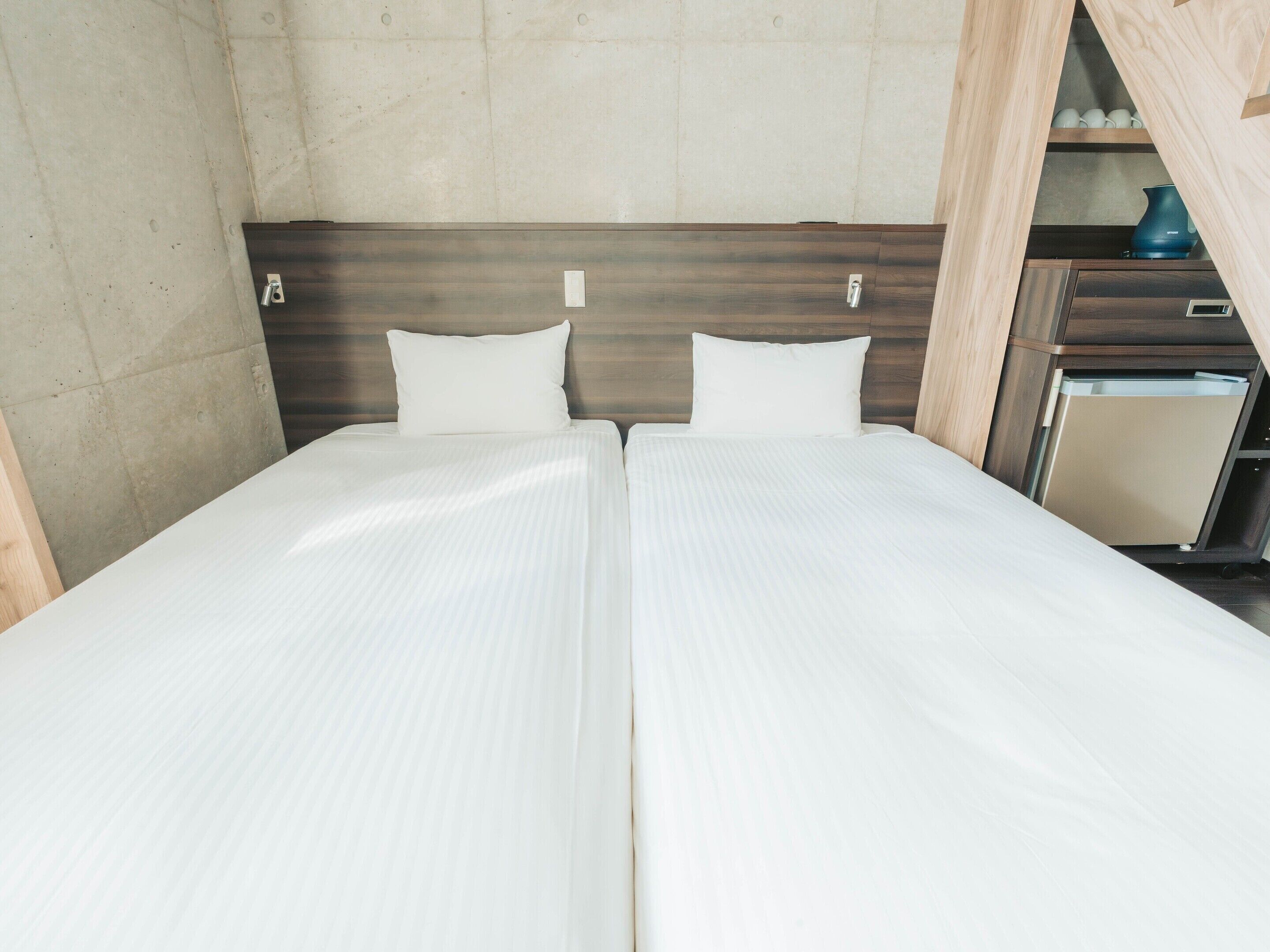 Una cama doble con cabecera de madera, una mesita de noche pequeña y una repisa empotrada con tazas y una tetera.