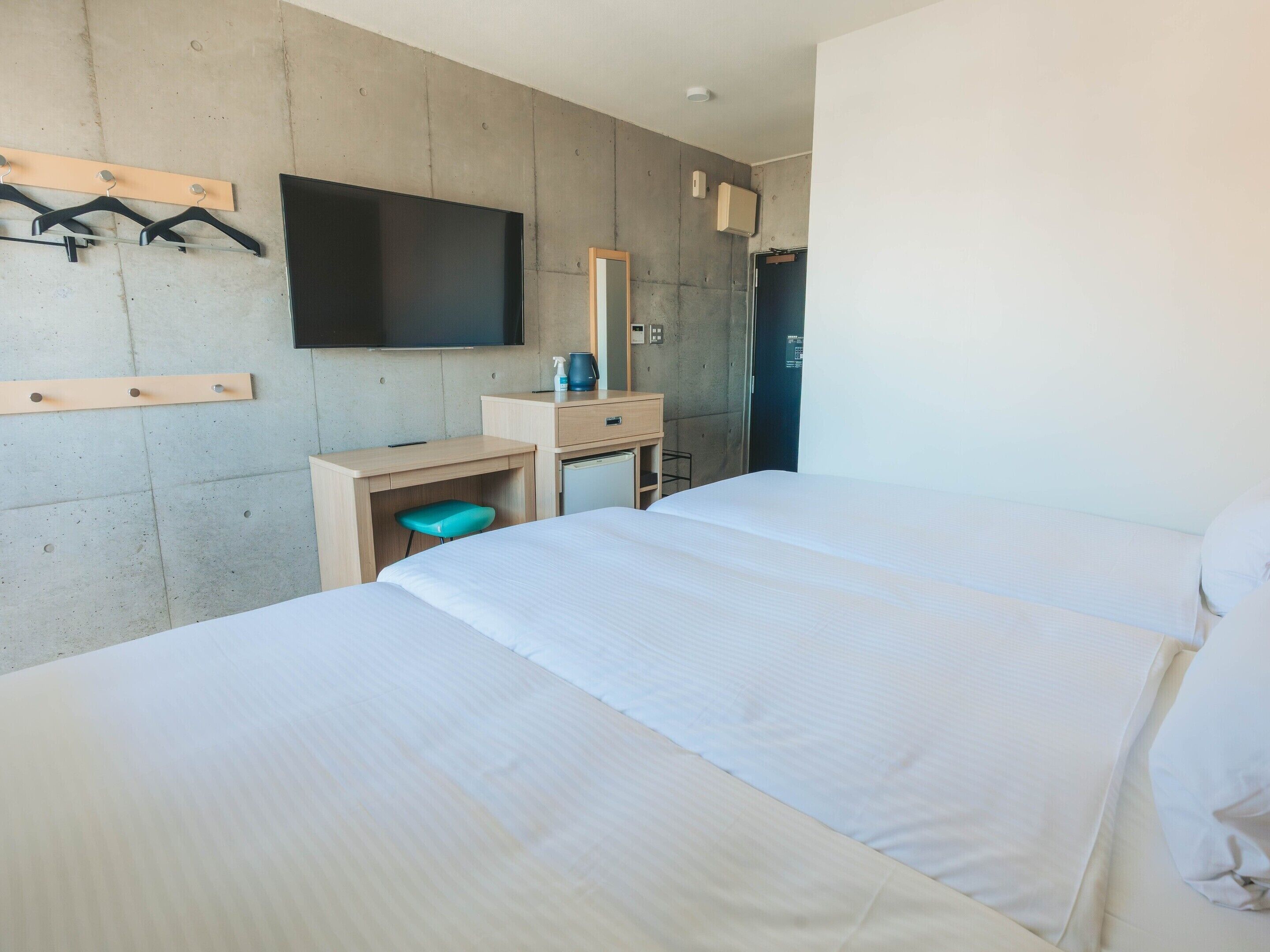 A hotel room with two beds, a desk with a chair, a television mounted on the wall, and a small kitchenette area.